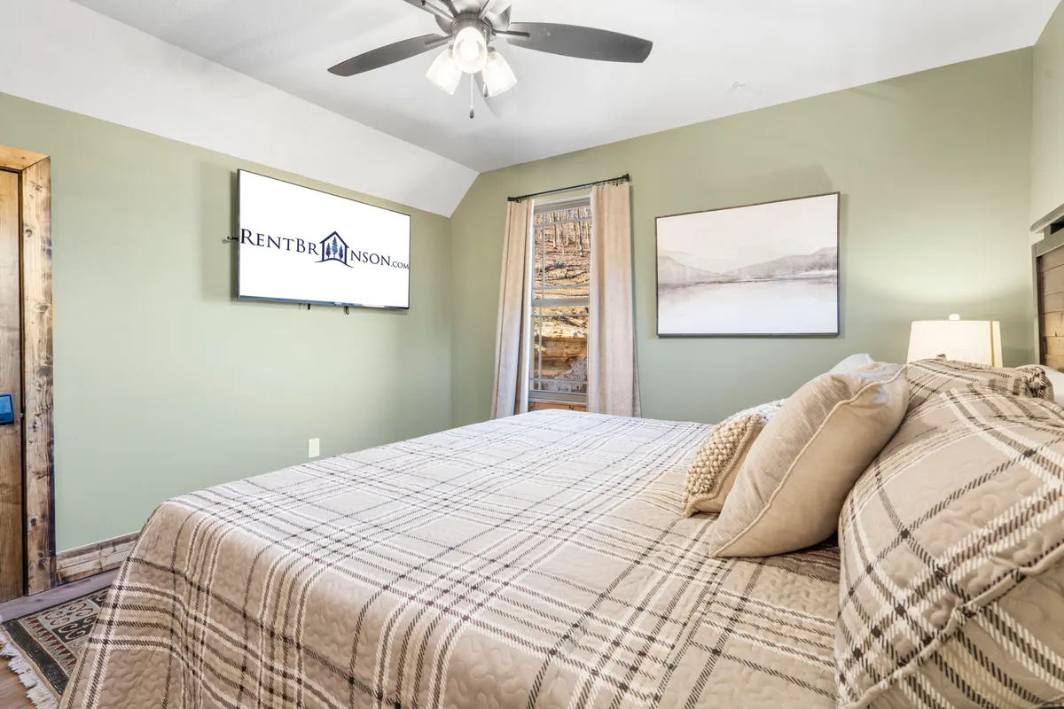 A cozy King bedroom showcasing a bed with plaid bedding, a mounted TV, curtains framing the window, and framed artwork on the wall. The room exudes warmth and offers a sense of privacy and relaxation, ideal for unwinding after a day of adventure.