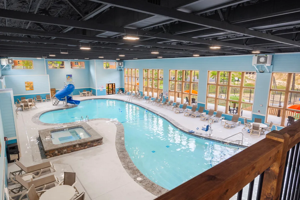 A bright, inviting indoor pool area with a swimming pool, water slide, hot tub, large windows letting in natural light, and lounge chairs. Families can be seen gathering, swimming, and relaxing poolside, capturing a mood of playful relaxation.