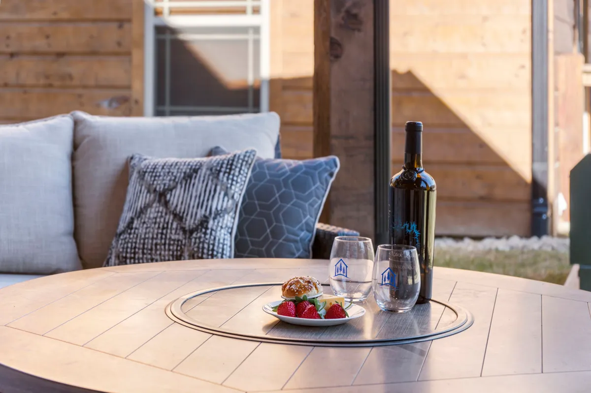 A close-up exterior shot of a small table set with a bottle of wine, glasses, a plate of strawberries and a croissant, and plush cushions, all atop a wooden deck or patio. Trees and greenery peek out in the soft background, suggesting a serene outdoor experience.