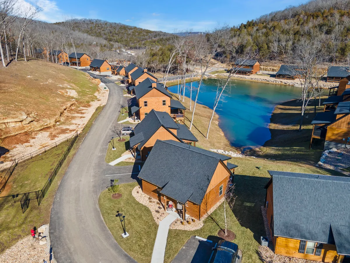 A tranquil exterior scene featuring cabins overlooking a serene pond, with a tree line and a winding pathway alongside the water. The area invites outdoor activities like walking, fishing, and relaxing by the lake, especially as the sun sets, creating a peaceful atmosphere.