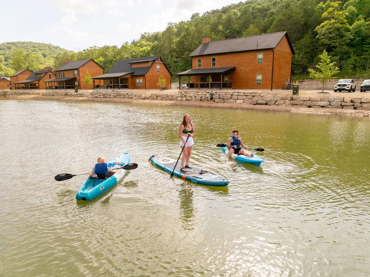 This image captures kayakers enjoying a sunny day on the water, with cozy cabins and a tranquil lakeside setting in the background. The water’s surface glistens, and the scene exudes a sense of outdoor adventure and family fun.