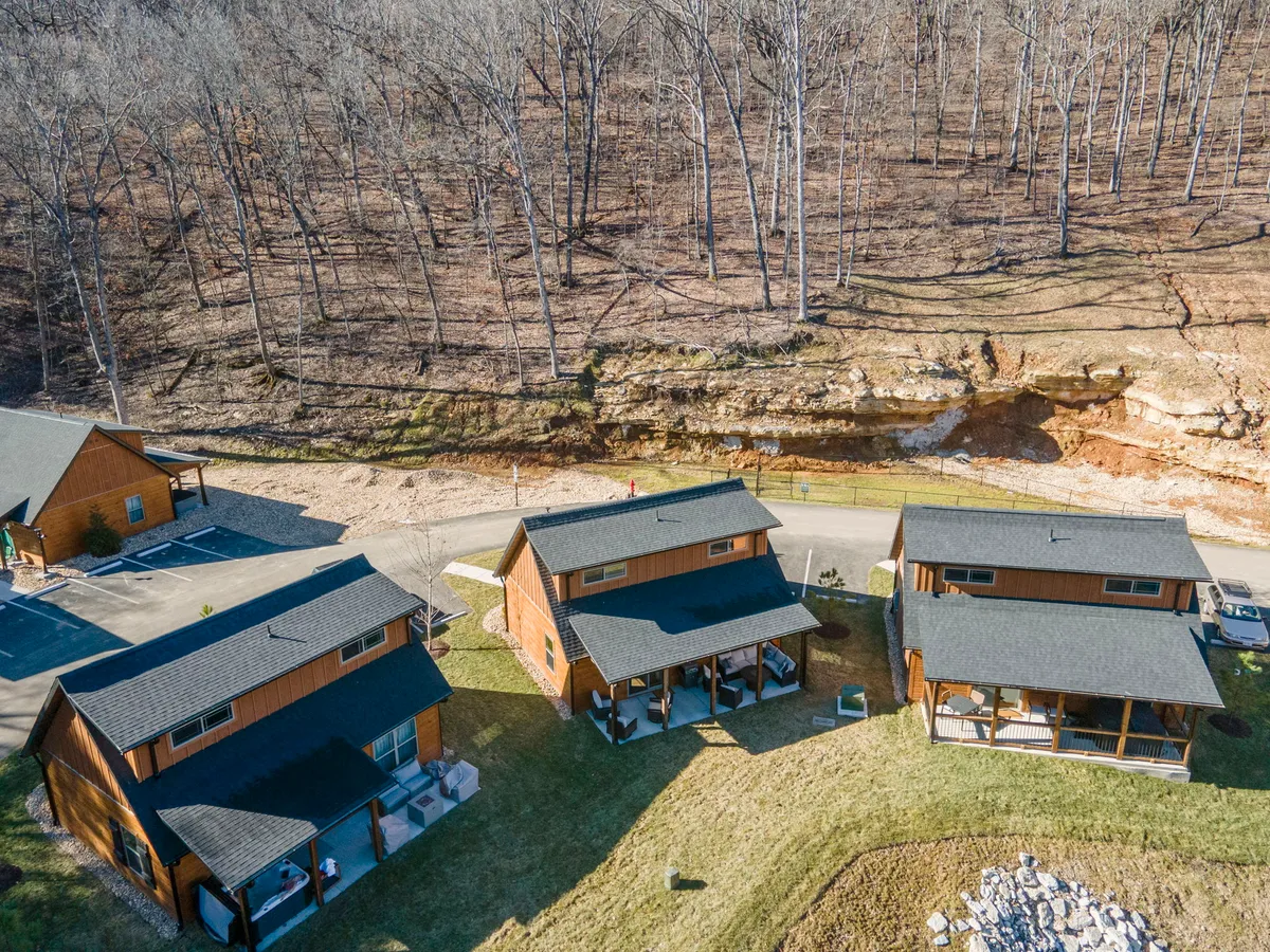 A tranquil exterior view of several wooden cabins set amid a green lawn, surrounded by mature trees and a natural rock formation. The porches face out onto the lawn, perfect for relaxing and taking in the peaceful Ozark scenery.
