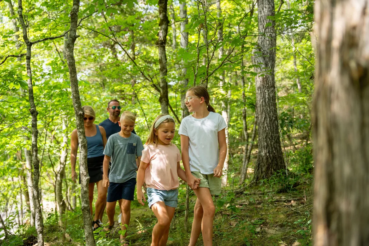 A family walks together along a leafy trail, sunlight streaming through the trees. The group appears joyful and carefree, immersed in nature and ready to explore the community’s scenic hiking paths.