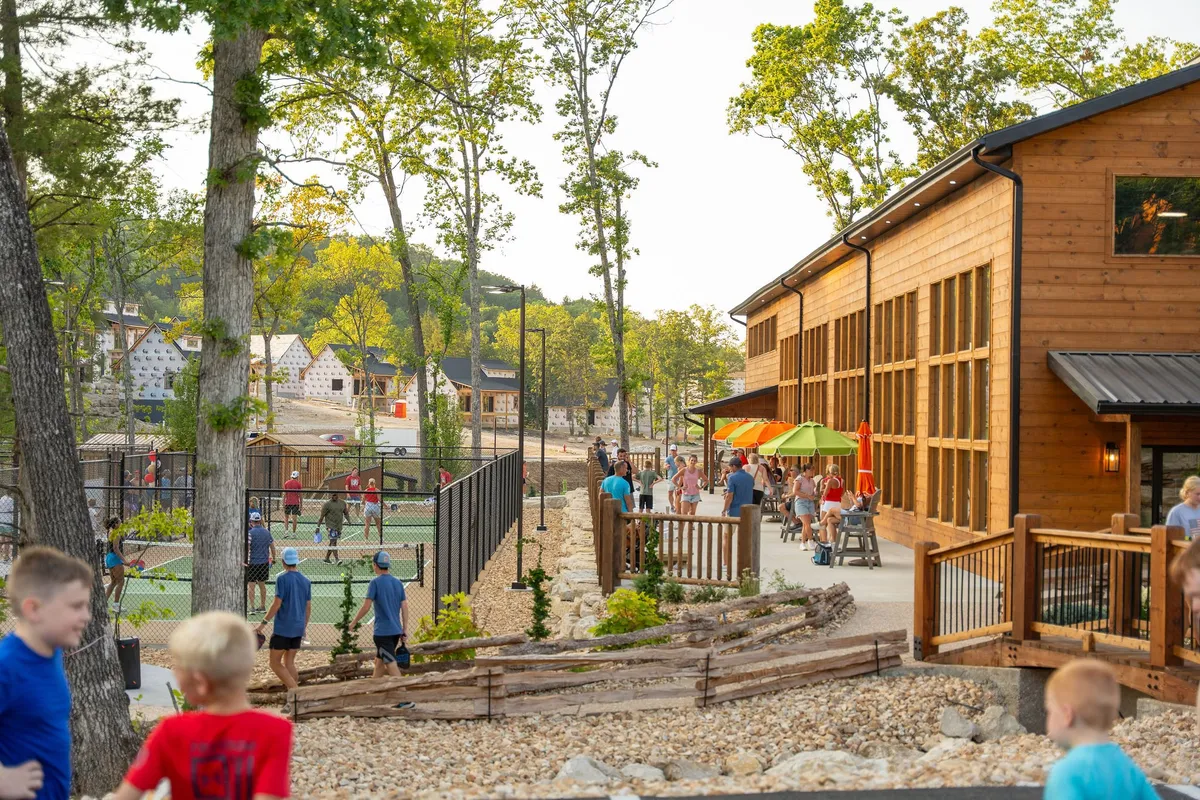 A lively exterior view showing a wooden building adjacent to tennis/pickleball courts, with children actively playing and families socializing at outdoor seating areas. The space is clearly designed for recreation, sports, and community gatherings.