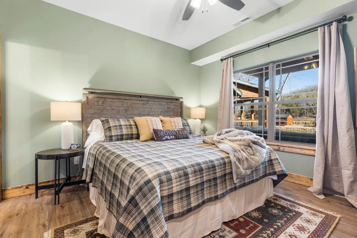 A warmly lit King bedroom featuring a plaid bedspread, wooden headboard, decorative pillows, a nightstand with a lamp, and a window dressed with curtains. The view from the window lets in natural light, contributing to the inviting and restful feel of the space. This room is styled for relaxation and comfort.