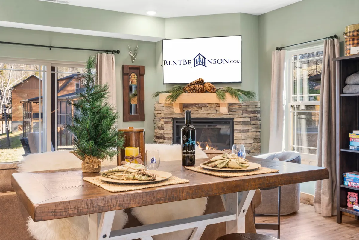 A cozy dining area centered on a wooden table set with decorative plates and a bottle of wine, with a fireplace in the background and a television for added comfort. The scene is warm and inviting, ideal for shared meals and relaxation.