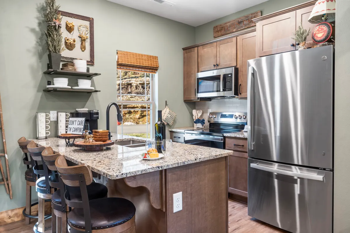 A warmly lit kitchen showcasing granite countertops, stainless steel appliances, and bar stools positioned at the counter. There is a window view letting in natural light, and the space is arranged for both cooking and casual dining or gathering.